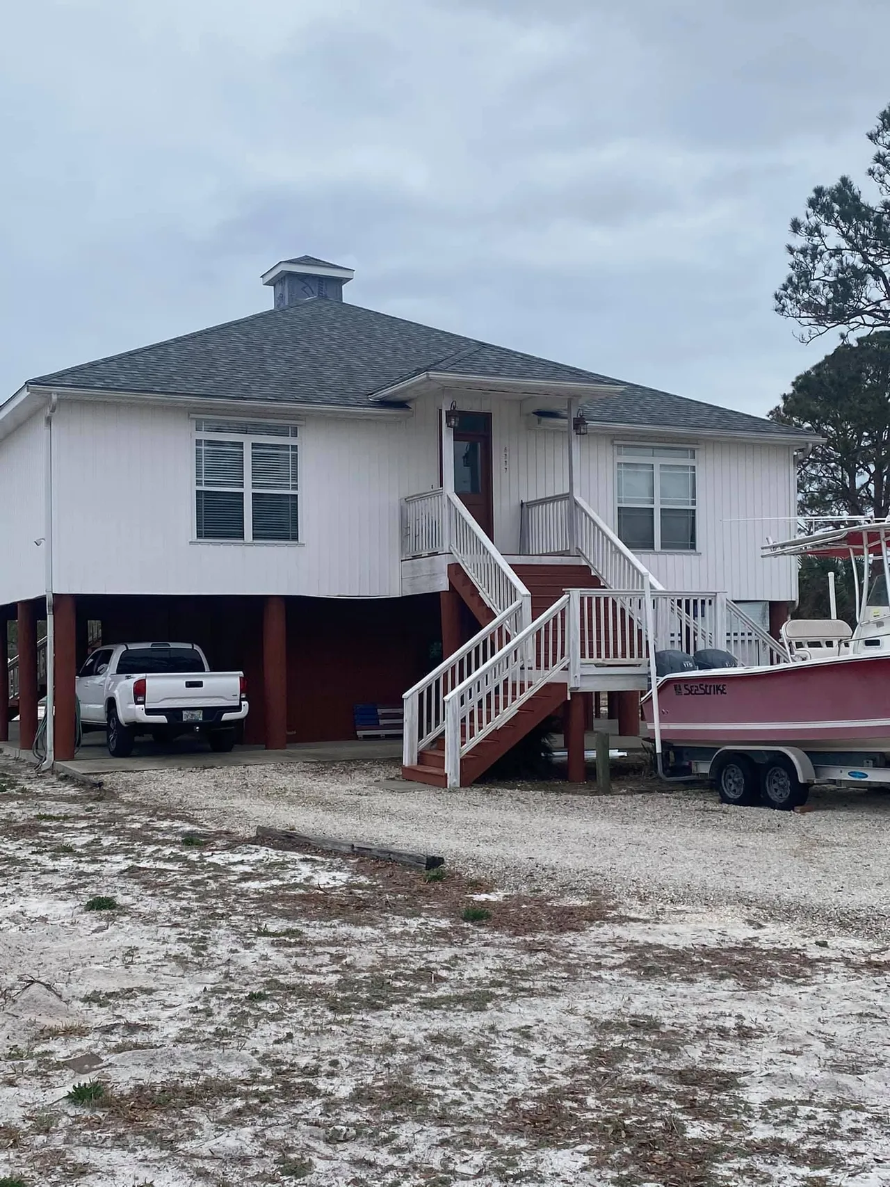 White house on stilts with a truck parked underneath. Boat on a trailer beside it. Cloudy day.