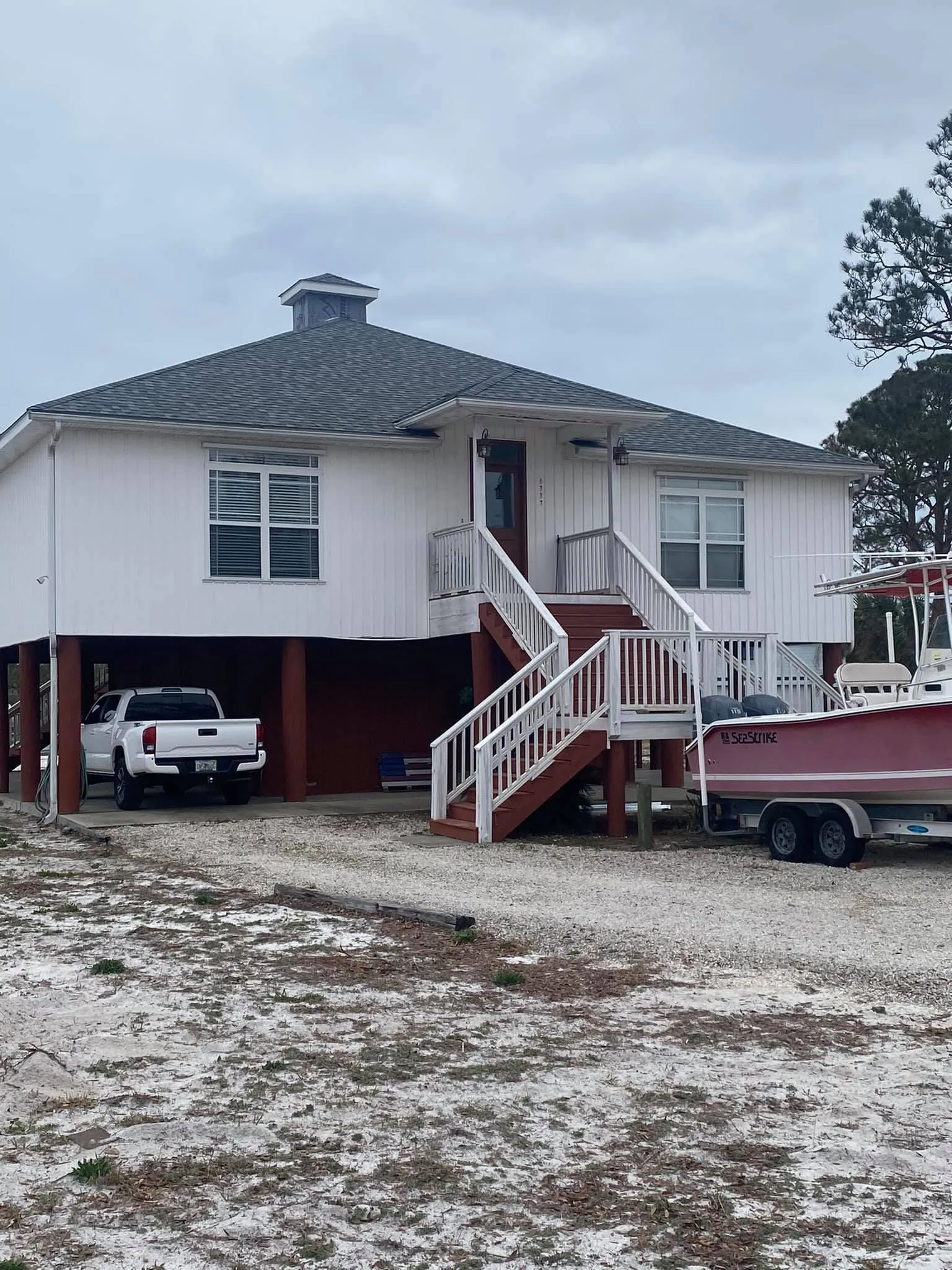 White house on stilts with a truck parked underneath. Boat on a trailer beside it. Cloudy day.