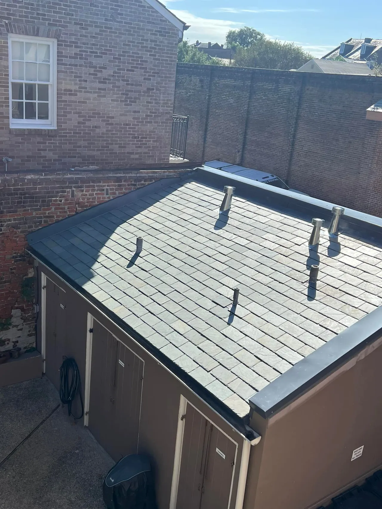 Brown shed with a dark roof in a brick courtyard; a window is visible.