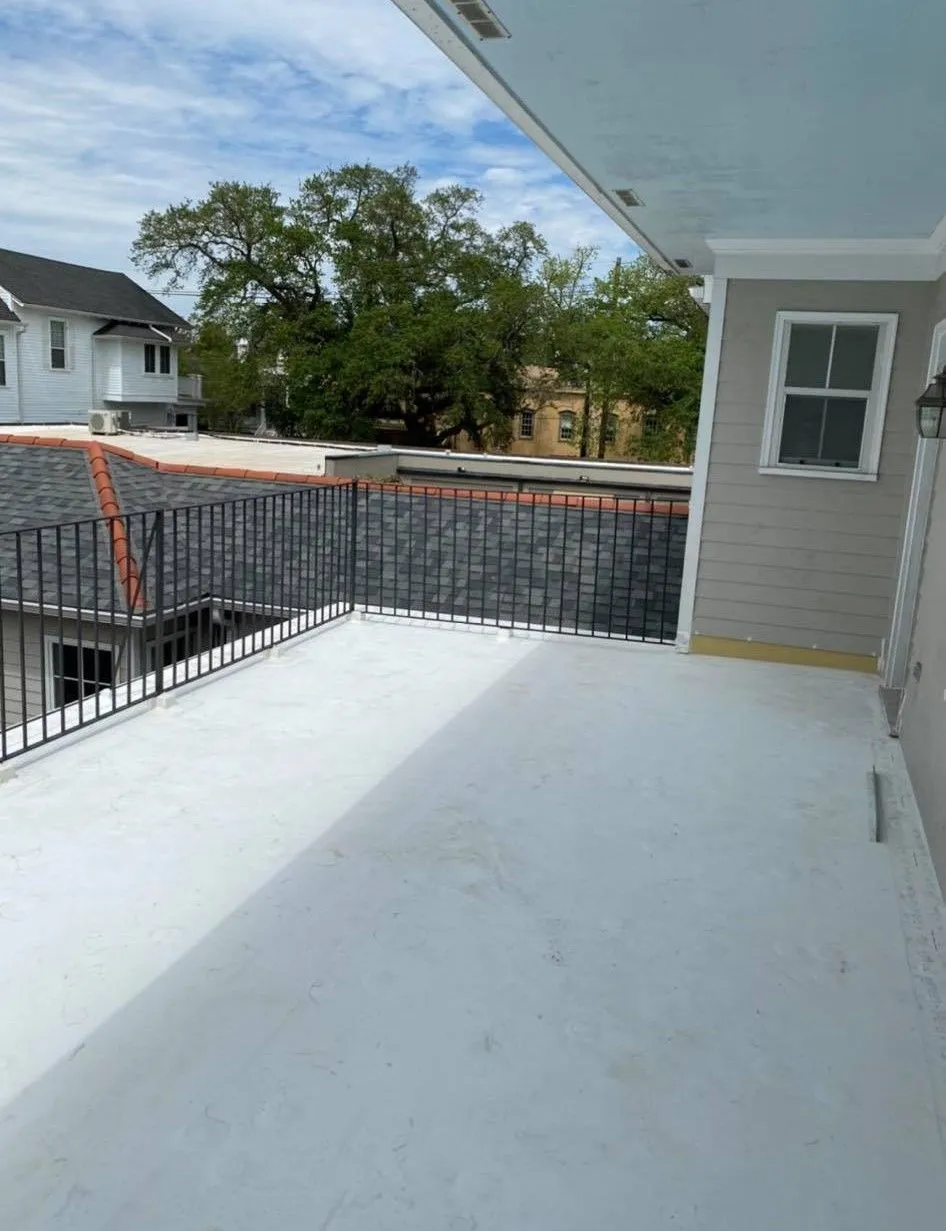 Rooftop deck with white surface, black railing, and grey siding. Buildings and trees in the background.