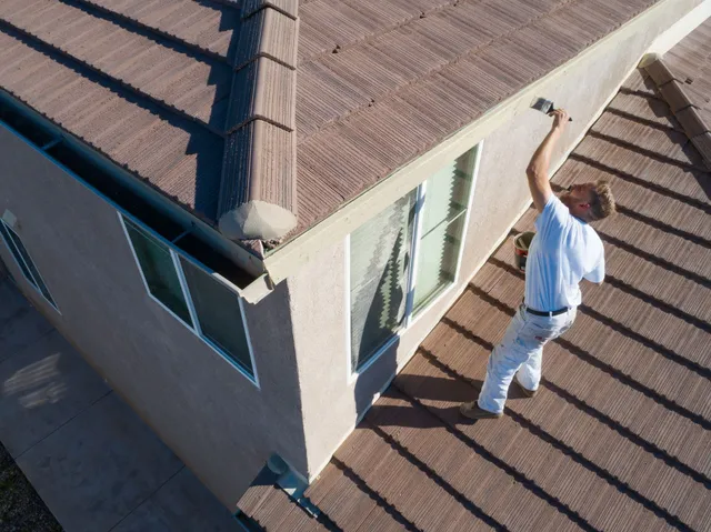 Person painting the edge of a tan house roof. The roof is brown.