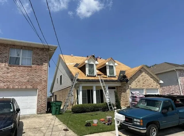 House under construction with workers on the roof. Ladders, a truck, and materials are in the yard.