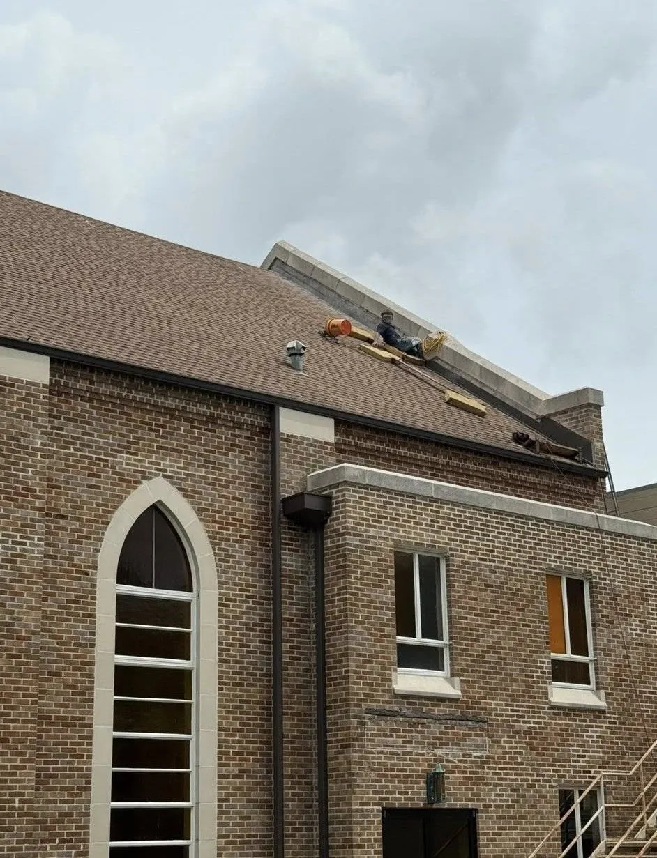 Brick church exterior with roof repairs in progress, brown shingles, cloudy sky.