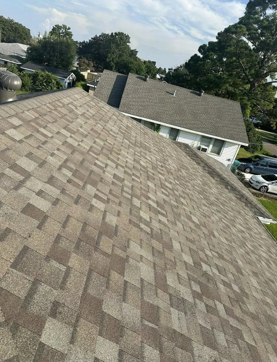 Overhead view of a roof with brown and gray asphalt shingles. Another roof and houses are in the background.