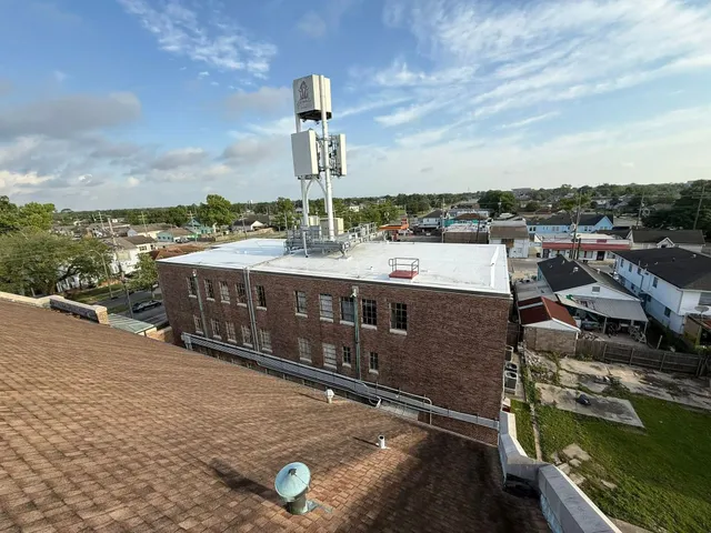 A brick building with a cell tower on the roof, surrounded by other buildings and a partly cloudy sky.