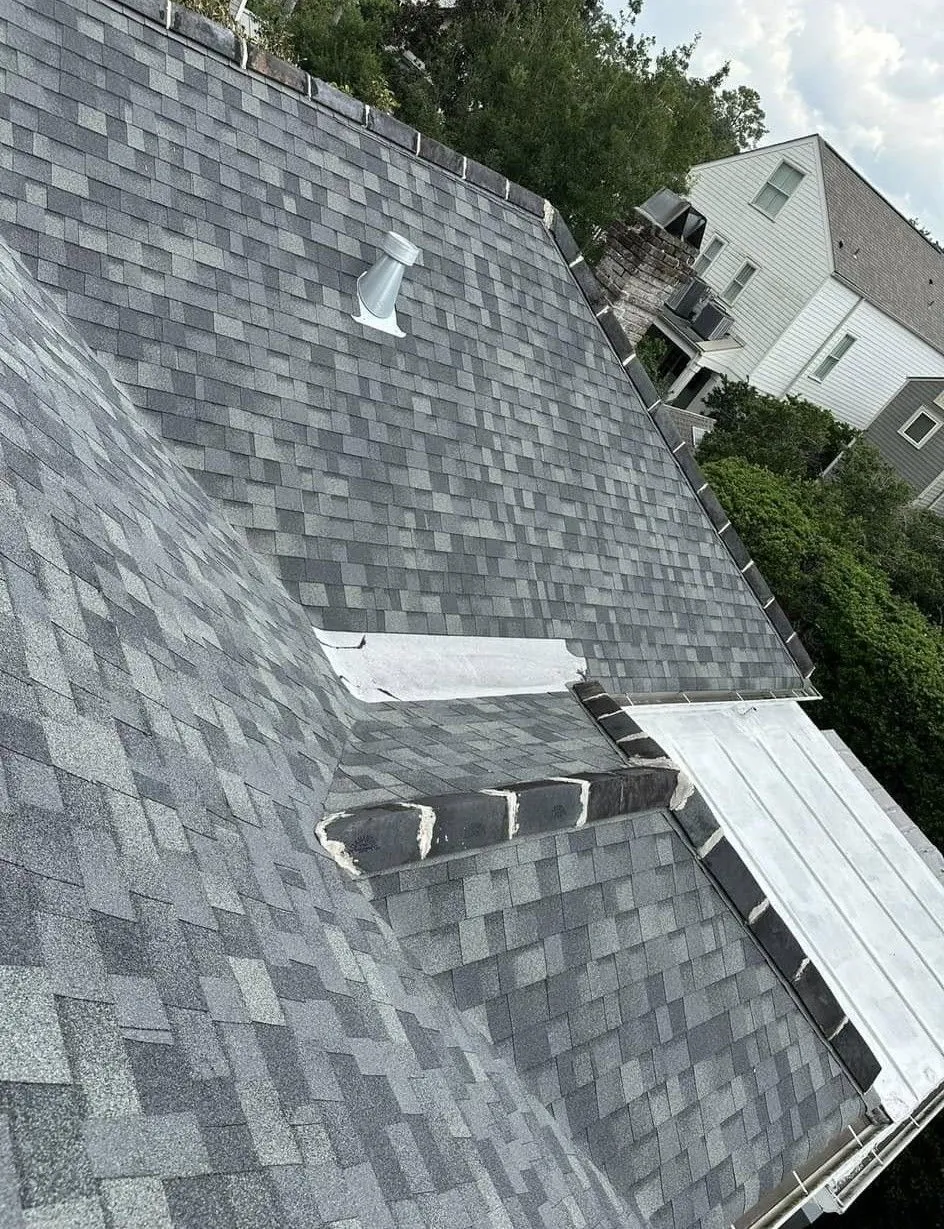 Gray asphalt shingle roof with a vent and a flat section. A house is in the background.